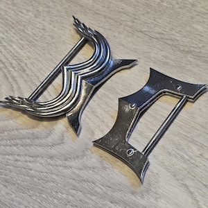 May include: Two silver-colored metal belt buckles with a decorative design. One buckle has an ornate, curved shape with a central bar. The other buckle is a more angular design with a straight bar. Both are on a wooden surface.