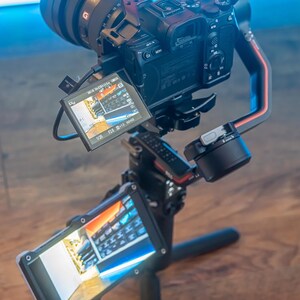 May include: A black camera with a large lens is mounted on a black and red camera stabiliser. The camera is pointed towards the ground and the screen is showing a preview of the video being recorded. The stabiliser is mounted on a black tripod with three legs.