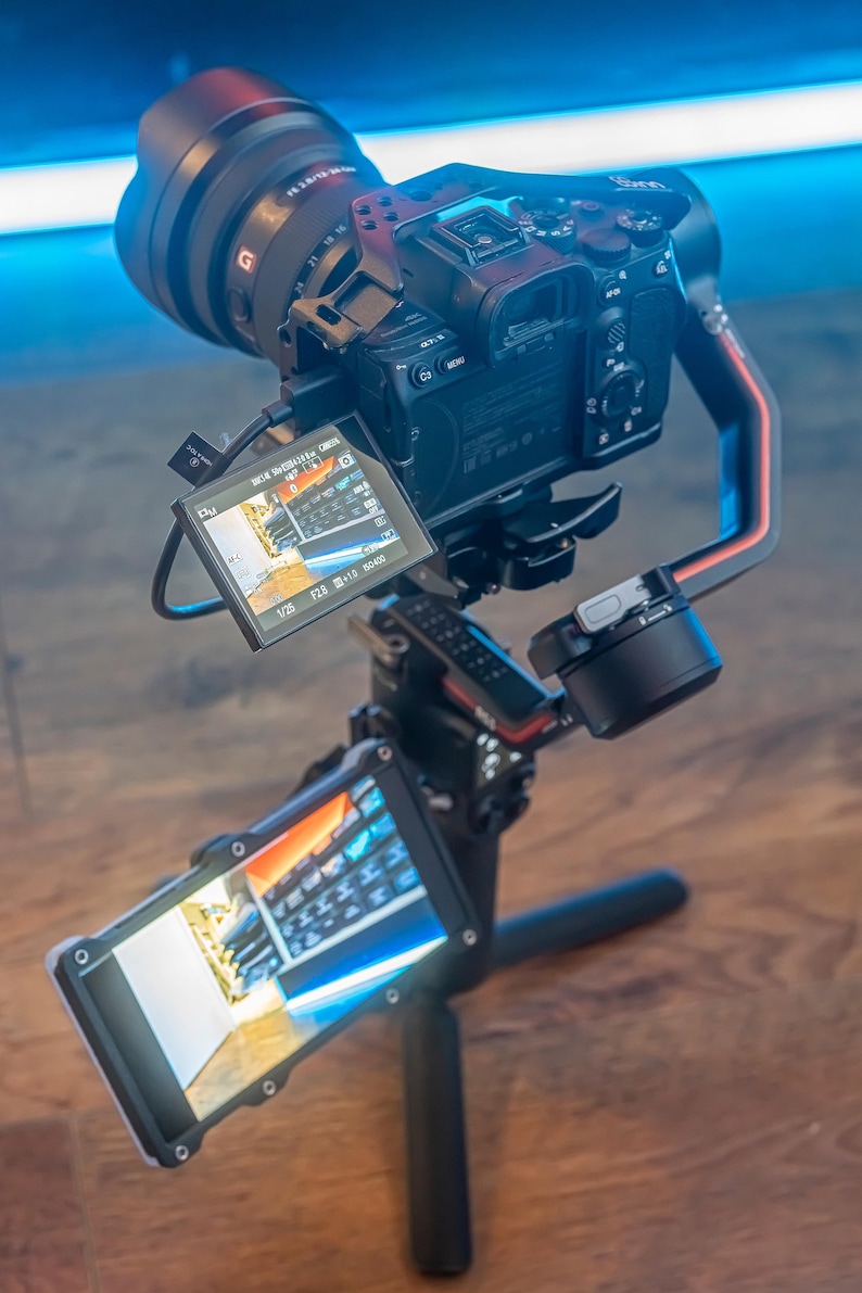 May include: A black camera with a large lens is mounted on a black and red camera stabiliser. The camera is pointed towards the ground and the screen is showing a preview of the video being recorded. The stabiliser is mounted on a black tripod with three legs.