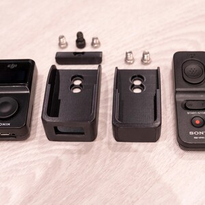Puede incluir: Una colecci&oacute;n de accesorios electr&oacute;nicos negros, que incluyen un dispositivo etiquetado como "RONIN", dos soportes de montaje y un control remoto Sony. Tambi&eacute;n hay peque&ntilde;as piezas de hardware plateadas y negras. Los art&iacute;culos est&aacute;n dispuestos sobre una superficie clara.