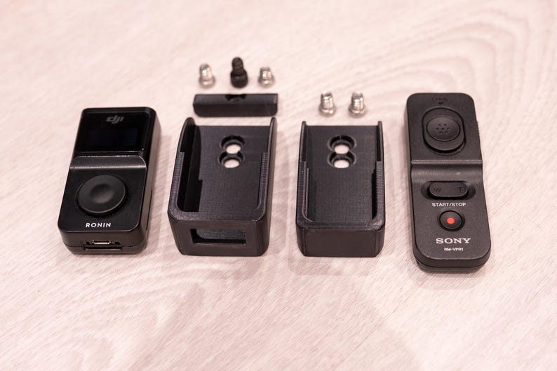 Puede incluir: Una colecci&oacute;n de accesorios electr&oacute;nicos negros, que incluyen un dispositivo etiquetado como "RONIN", dos soportes de montaje y un control remoto Sony. Tambi&eacute;n hay peque&ntilde;as piezas de hardware plateadas y negras. Los art&iacute;culos est&aacute;n dispuestos sobre una superficie clara.
