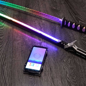 Pode incluir: Duas espadas luminosas, uma com cabo preto e l&acirc;mina arco-&iacute;ris, a outra com cabo preto e prateado e l&acirc;mina roxa. Um smartphone com um aplicativo de mudan&ccedil;a de cor est&aacute; em primeiro plano.