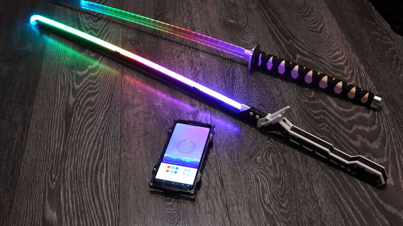 Pode incluir: Duas espadas luminosas, uma com cabo preto e l&acirc;mina arco-&iacute;ris, a outra com cabo preto e prateado e l&acirc;mina roxa. Um smartphone com um aplicativo de mudan&ccedil;a de cor est&aacute; em primeiro plano.