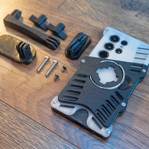May include: A black and silver 3D printed phone case with a circular cutout and a ring holder. The case is disassembled and laid out on a wooden surface with screws and other parts.