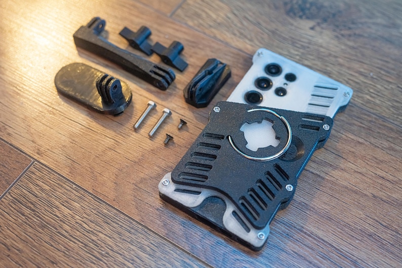 May include: A black and silver 3D printed phone case with a circular cutout and a ring holder. The case is disassembled and laid out on a wooden surface with screws and other parts.