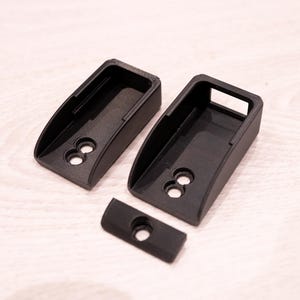 Puede incluir: Tres componentes de pl&aacute;stico negro impresos en 3D. Dos piezas m&aacute;s grandes tienen forma rectangular con la parte superior curvada y dos agujeros circulares. Tambi&eacute;n hay una pieza m&aacute;s peque&ntilde;a, rectangular, con un solo agujero.