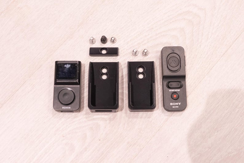 Puede incluir: Un conjunto de dispositivos y accesorios electr&oacute;nicos negros y grises, que incluyen un peque&ntilde;o dispositivo con pantalla etiquetado como "RONIN", dos fundas protectoras negras, un mando a distancia "SONY" y varios tornillos.