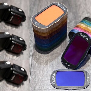 May include: Three flash units with different colored gels attached. The gels are purple, blue, and yellow. The flash units are black with a red button on the side. The gels are labeled with the color and a +1.