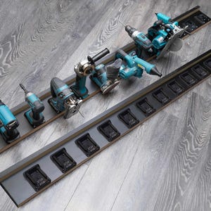 May include: A collection of teal and black power tools, including a drill, saw, and grinder, arranged on two gray wooden storage racks. The racks have slots for tool batteries. The tools are displayed on a gray wooden surface.