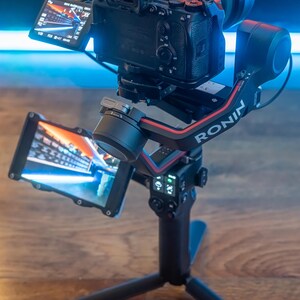 May include: A black camera with a lens attached to a black and red camera stabiliser with the word "RONIN" printed on the side. The camera is mounted on a black tripod with three legs.