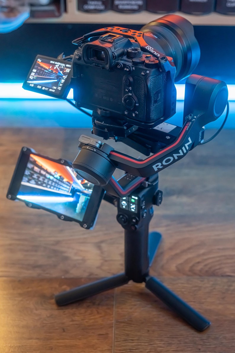 May include: A black camera with a lens attached to a black and red camera stabiliser with the word "RONIN" printed on the side. The camera is mounted on a black tripod with three legs.