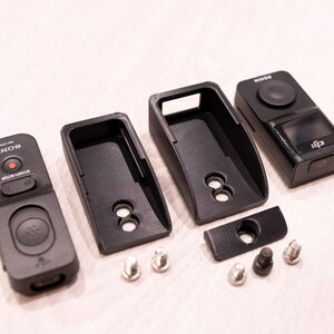 Puede incluir: Un conjunto de dispositivos electr&oacute;nicos y accesorios negros, que incluyen un mando a distancia con el logotipo "SONY", dos fundas protectoras, una c&aacute;mara y varios tornillos peque&ntilde;os. Los art&iacute;culos est&aacute;n dispuestos sobre una superficie clara.
