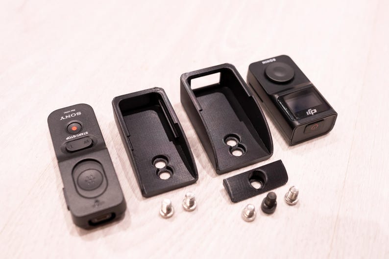 Puede incluir: Un conjunto de dispositivos electr&oacute;nicos y accesorios negros, que incluyen un mando a distancia con el logotipo "SONY", dos fundas protectoras, una c&aacute;mara y varios tornillos peque&ntilde;os. Los art&iacute;culos est&aacute;n dispuestos sobre una superficie clara.