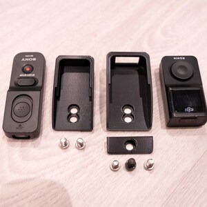 Puede incluir: Accesorios de c&aacute;mara negros, que incluyen un control remoto etiquetado como "SONY", dos soportes de montaje y un controlador etiquetado como "RONIN". Tambi&eacute;n se incluyen varios tornillos peque&ntilde;os plateados y negros. Los art&iacute;culos est&aacute;n dispuestos sobre una superficie de madera clara.