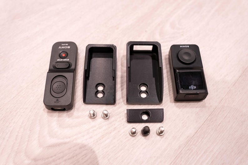 Puede incluir: Accesorios de c&aacute;mara negros, que incluyen un control remoto etiquetado como "SONY", dos soportes de montaje y un controlador etiquetado como "RONIN". Tambi&eacute;n se incluyen varios tornillos peque&ntilde;os plateados y negros. Los art&iacute;culos est&aacute;n dispuestos sobre una superficie de madera clara.