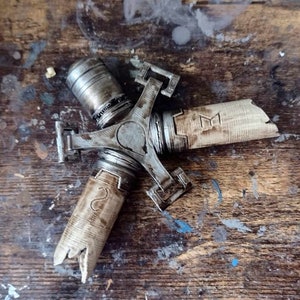 May include: A three-pronged metal object with a weathered, aged finish. Each prong has a wooden handle with a worn, distressed look. The metal prongs are connected by a central hub with a circular design. The wooden handles have markings that appear to be letters or symbols.