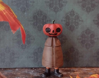 Könnte beinhalten: Eine kleine, braune Figur mit einem Kürbis-Kopf. Der Kürbis ist orange und hat ein geschnitztes Jack-o'-Lantern-Gesicht. Die Figur trägt einen braunen Mantel und schwarze Schuhe. Die Figur steht auf einer Holzoberfläche.