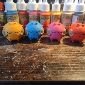 Può includere: Quattro figurine sferiche colorate, con corna e occhi chiusi, sono disposte su una superficie di legno. Le figurine sono dipinte in arancione, blu, rosa e rosso. Sullo sfondo sono visibili barattoli di vernice.
