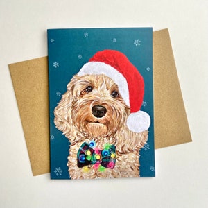 May include: A Christmas card featuring a golden doodle dog wearing a red Santa hat and a colourful bow tie against a teal background with white snowflakes.