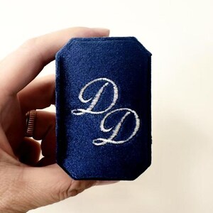 May include: A navy blue velvet ring box with white embroidered "DD" initials. The box is held in a hand, showcasing its octagonal shape and soft texture. A simple and elegant design.