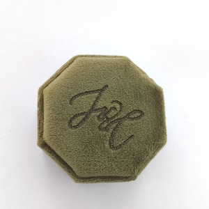 May include: An octagonal ring box covered in olive green velvet fabric. The top lid features a dark brown embroidered script monogram that appears to read J and L, centered on the soft, textured surface of the closed jewelry container.