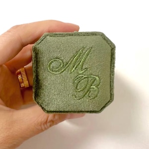 May include: Olive green velvet ring box with embroidered initials "MB". The box has an octagonal shape and a soft, textured appearance. A gold ring is visible in the background.