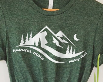 May include: Green t-shirt with a white mountain graphic, trees, and the text "wander more worry less." The shirt is hanging on a wooden hanger. A beige hat is visible in the background.