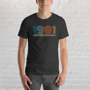 May include: A dark heather grey t-shirt with a vintage-style graphic of the year 1981 in colorful stripes. The text "LIMITED EDITION" is printed below the year.