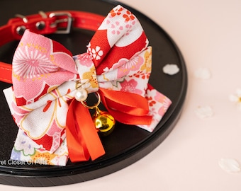 May include: Red leather pet collar with a floral bow tie. The bow tie features a red and white floral pattern, a gold bell, and a pearl charm. The collar is on a black surface with a pink background.