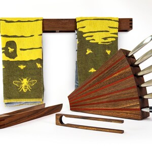 May include: A set of kitchen tools made of wood with red accents. The set includes a knife block with a fan-shaped design, a towel rack, and two pairs of tongs. The knife block holds a set of silver knives. The towels are yellow and green with a bee design.