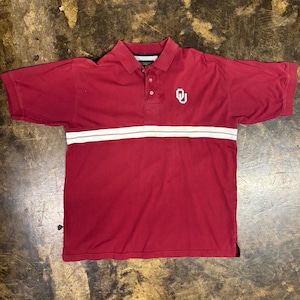 May include: A maroon short-sleeve polo shirt featuring a white horizontal stripe across the chest and a white OU logo embroidered on the upper left side. The shirt has a classic collar, a three-button placket, and ribbed sleeve cuffs.