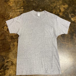 May include: A short-sleeved heather gray t-shirt featuring a crew neckline and a single patch pocket on the left side of the chest. The garment is constructed from a speckled knit fabric and includes a small white brand label at the inner collar.