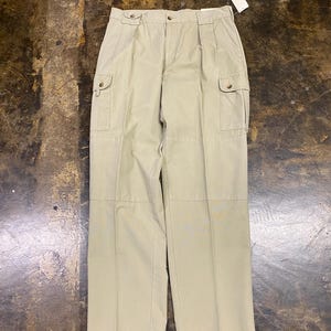 May include: Beige cargo pants featuring a button-front closure, belt loops, and two side cargo pockets with buttoned flaps. The trousers have a straight-leg cut with visible vertical seams running down the front of each leg and a small white tag.