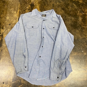 May include: A light blue long-sleeve button-down shirt featuring a pointed collar, two chest pockets with button closures, and dark buttons down the front placket. The shirt has a textured fabric appearance and buttoned cuffs on each sleeve.