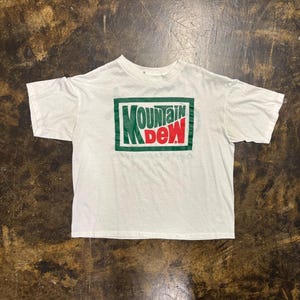 May include: A white short-sleeve t-shirt featuring a rectangular graphic on the chest. The graphic contains the text Mountain Dew in green and red lettering, enclosed within a green border. The shirt has a crew neckline and a standard fit.