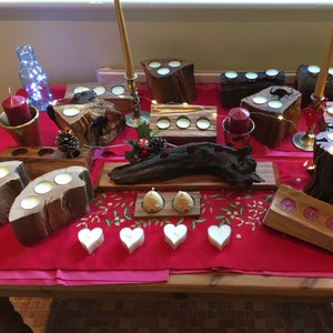 Puede incluir: Una colección de candelabros de madera con varios diseños, incluyendo un tronco, un tocón de árbol y una rama. Los candelabros están dispuestos sobre un mantel rojo con luces blancas y pequeñas velas blancas.