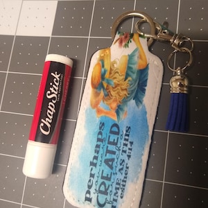 May include: A white fabric keychain with a blue tassel. The keychain features a watercolor-style image of a woman with a quote that reads "perhaps you were created for such a time as this esther 4:14".