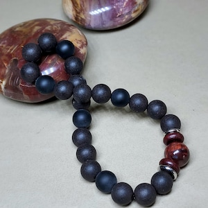 May include: A black beaded bracelet with a dark wood bead accent. The bracelet is made of round beads and is lying on a light brown surface.