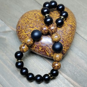 May include: Two beaded bracelets with black and brown wooden beads and gold accents. The bracelets are on a heart-shaped stone with brown and gold markings.