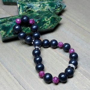 May include: A beaded bracelet featuring dark gray, spherical beads interspersed with faceted, deep pink beads. The bracelet has silver-toned accents and is displayed on a wooden surface, with two green crystal formations in the background.
