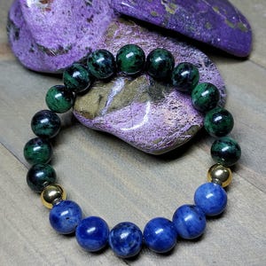 May include: A beaded bracelet featuring a combination of green, blue, and gold beads. The bracelet is made with round beads in shades of green, deep blue, and gold. The bracelet is displayed on a wooden surface with purple stones in the background.