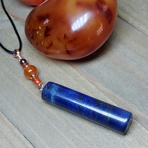 May include: A handcrafted necklace featuring a dark blue lapis lazuli pendant with copper accents and a black cord. The necklace includes orange and brown beads. Two polished orange stones are in the background. The necklace is displayed on a wooden surface.