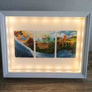 May include: A white framed artwork with three panels, each depicting a different Pokémon character in a colorful outdoor scene. The frame has built-in LED lights. The artwork is displayed on a wooden surface.