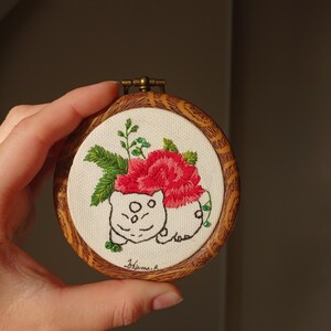 May include: A white embroidered design of a cartoon animal with a red rose and green leaves on a white canvas, framed in a round wooden frame.