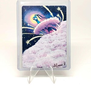 May include: A trading card featuring a blue and white creature emerging from a cloud, set against a starry night sky. The artwork includes a signature in the lower right corner and is encased in a protective sleeve.