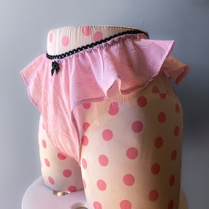 Pode incluir: Lingerie rosa com acabamento em renda preta e borda de babados. A lingerie está em um manequim de bolinhas rosa e branco.