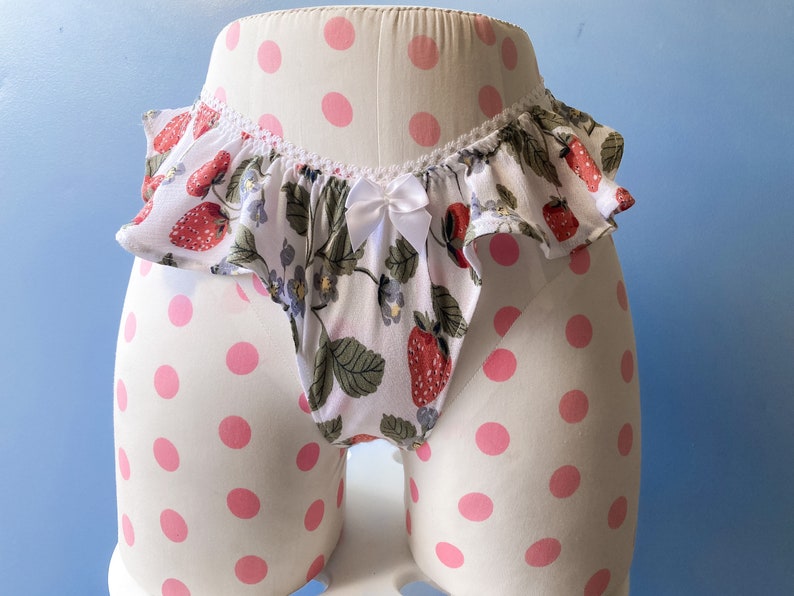 May include: White underwear with a ruffled top and a strawberry print. The underwear is on a white background with pink polka dots.