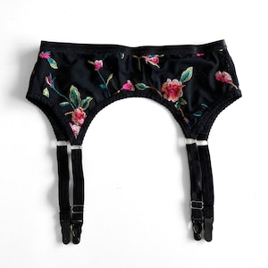 May include: Black garter belt with floral embroidery. The belt has two adjustable straps with metal clips.