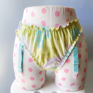 Puede incluir: Lingerie de estampado floral verde y blanco con ligas azules. La lencería está en un maniquí blanco con lunares rosas.