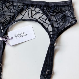 Puede incluir: Ligero de liga negro con un dise&ntilde;o de telara&ntilde;a. El liguero tiene dos correas ajustables con cierres plateados. La etiqueta dice "Reverie Intimates".